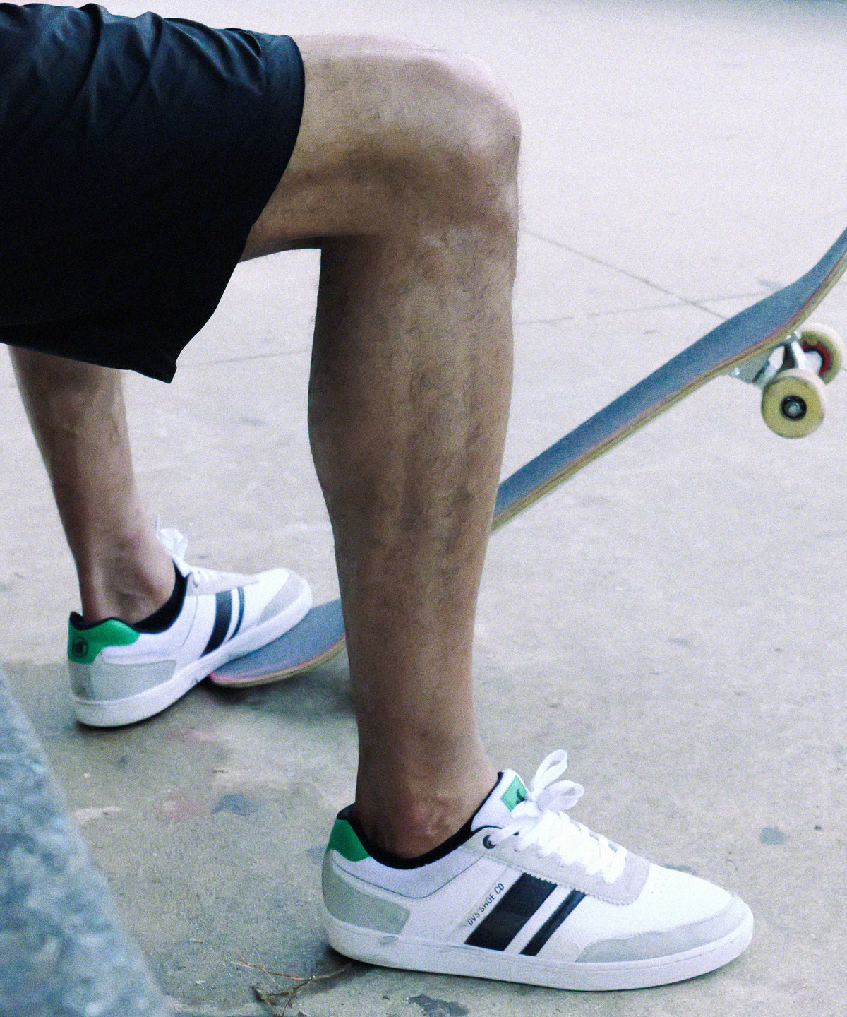 Person wearing white sneakers with green accents and black stripes, sitting on a skateboard.