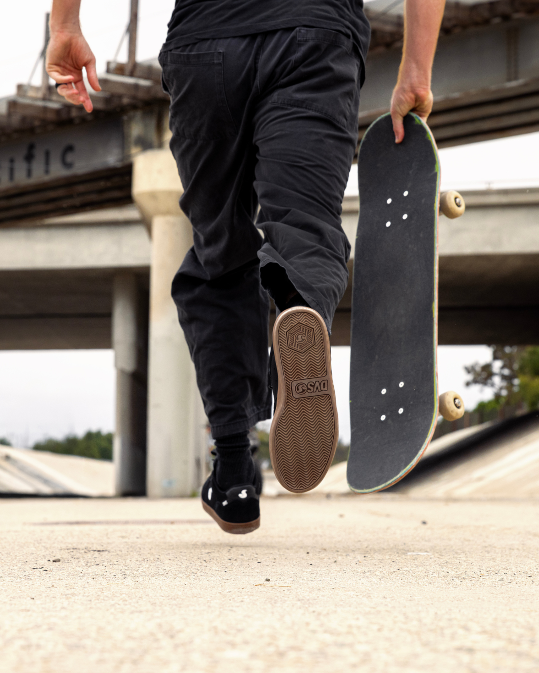 A person in black pants and black sneakers walks under a concrete bridge while holding a skateboard upside down in one hand. The skateboard deck is black with natural wood edges and beige wheels.