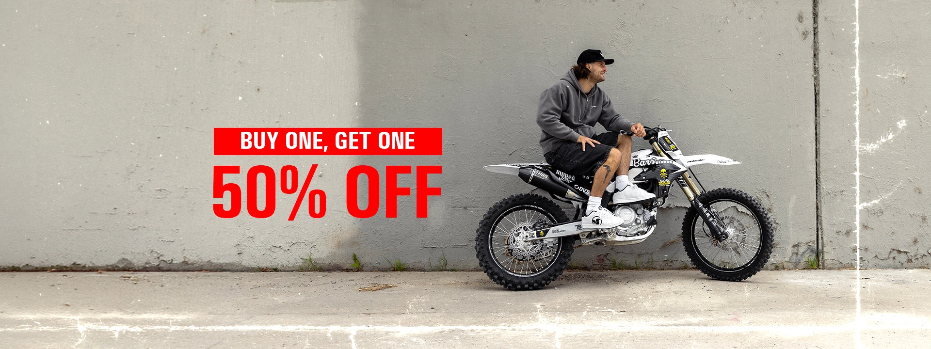 A man wearing a black cap, gray hoodie, and black shorts sits on a white and black off-road dirt bike against a gray concrete wall. A red promotional banner on the left reads 'BUY ONE, GET ONE 50% OFF'.