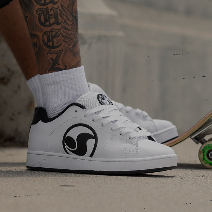 A person wearing white low-top sneakers with black graphic logos, standing on a concrete surface next to a skateboard. The image has a dark overlay and a black banner at the bottom reading 'SHOP REVIVAL'.