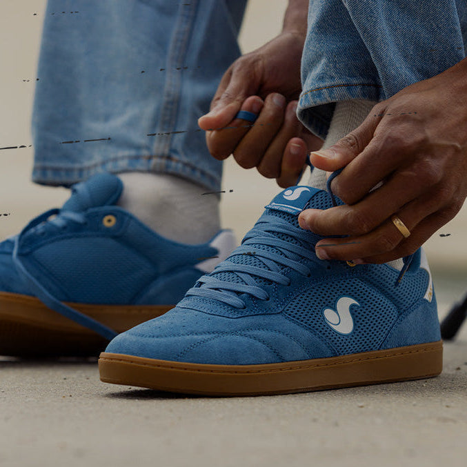 A person wearing blue suede and mesh Core 95 skate shoes with gum soles, sitting on a concrete surface. The person is tying the laces of the right shoe, with jeans and a gold ring visible.