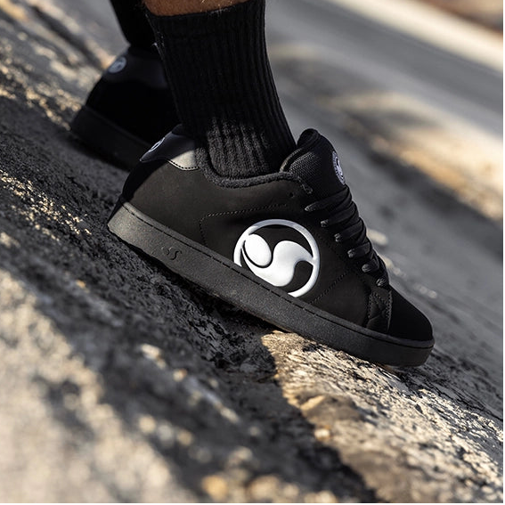 A close-up lifestyle shot of a person sitting outdoors on a concrete ledge, wearing black low-top sneakers with a white circular logo, black crew socks, and black denim shorts.