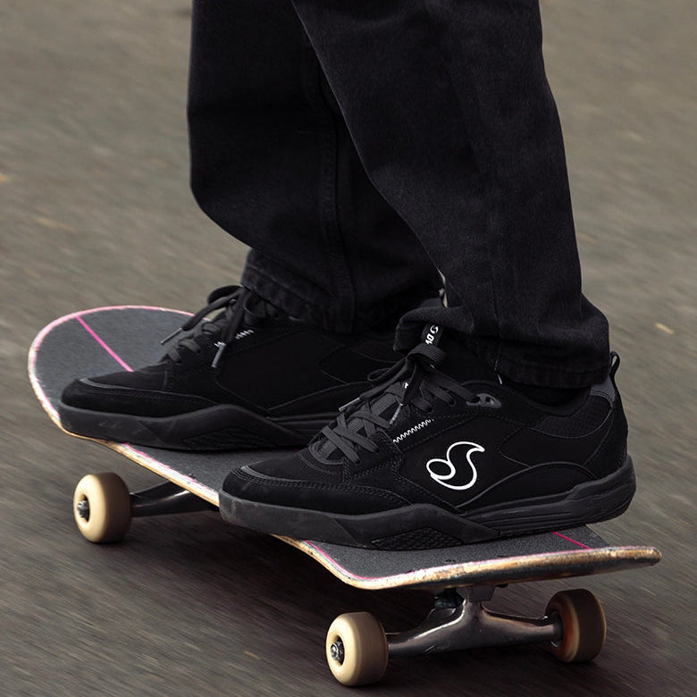 A close-up of a person's legs and feet wearing black skate shoes with a white logo, standing on a skateboard on a paved street.