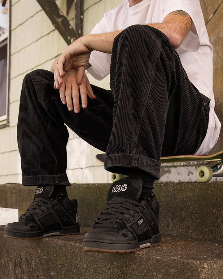 Person wearing black sneakers with visible branding, sitting on a step with a skateboard.