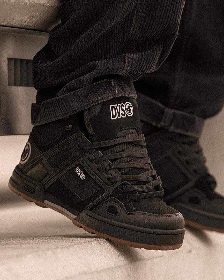 Black high-top sneakers with 'DVS' logo worn by a person on a neutral background