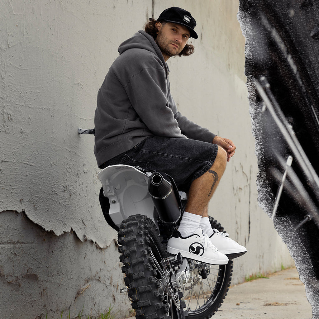 A man wearing a black cap, gray hoodie, and black shorts sits on a white dirt bike against a textured concrete wall.