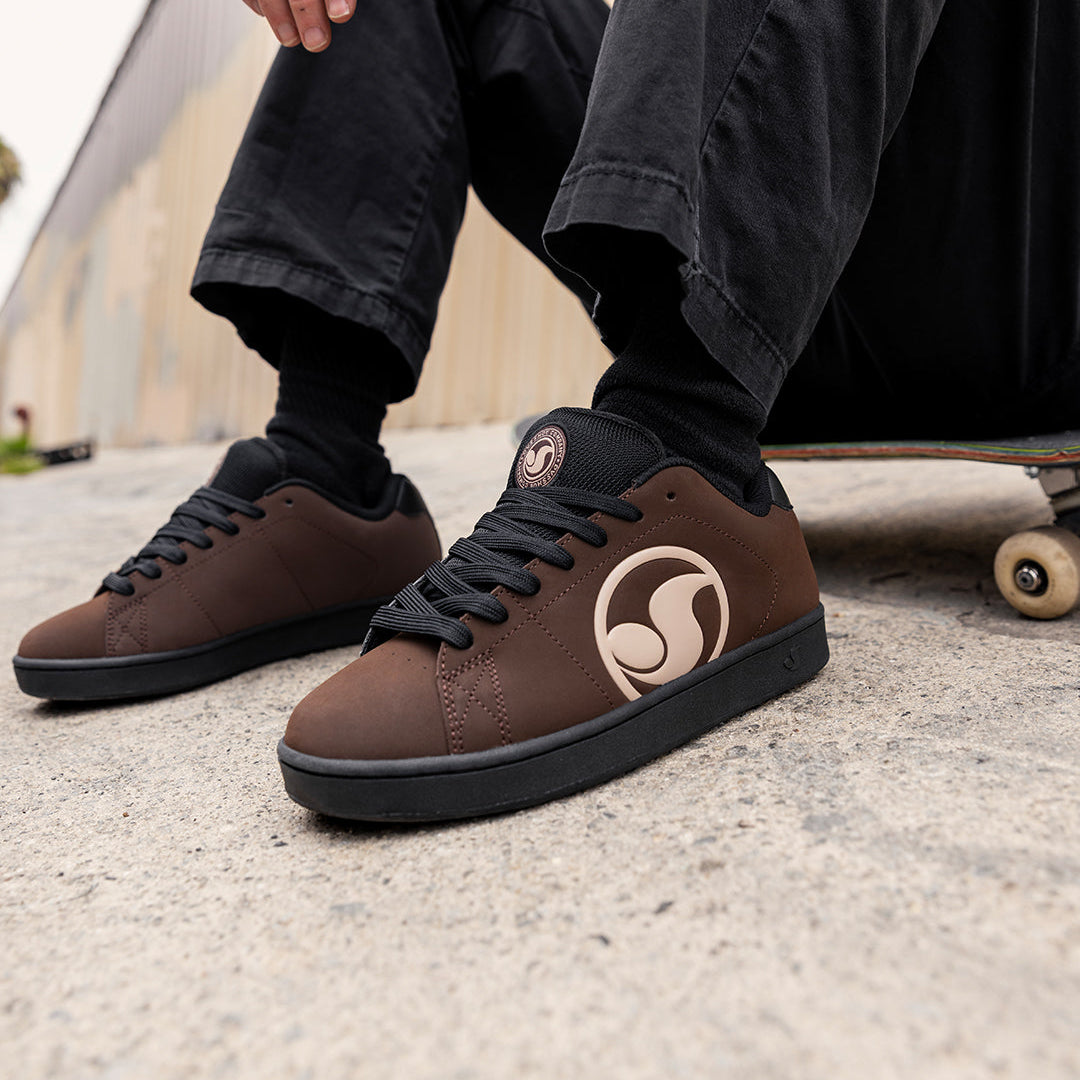 A person sitting on a skateboard outdoors wearing brown suede sneakers with black soles and a circular tan logo on the side, paired with black pants and socks.
