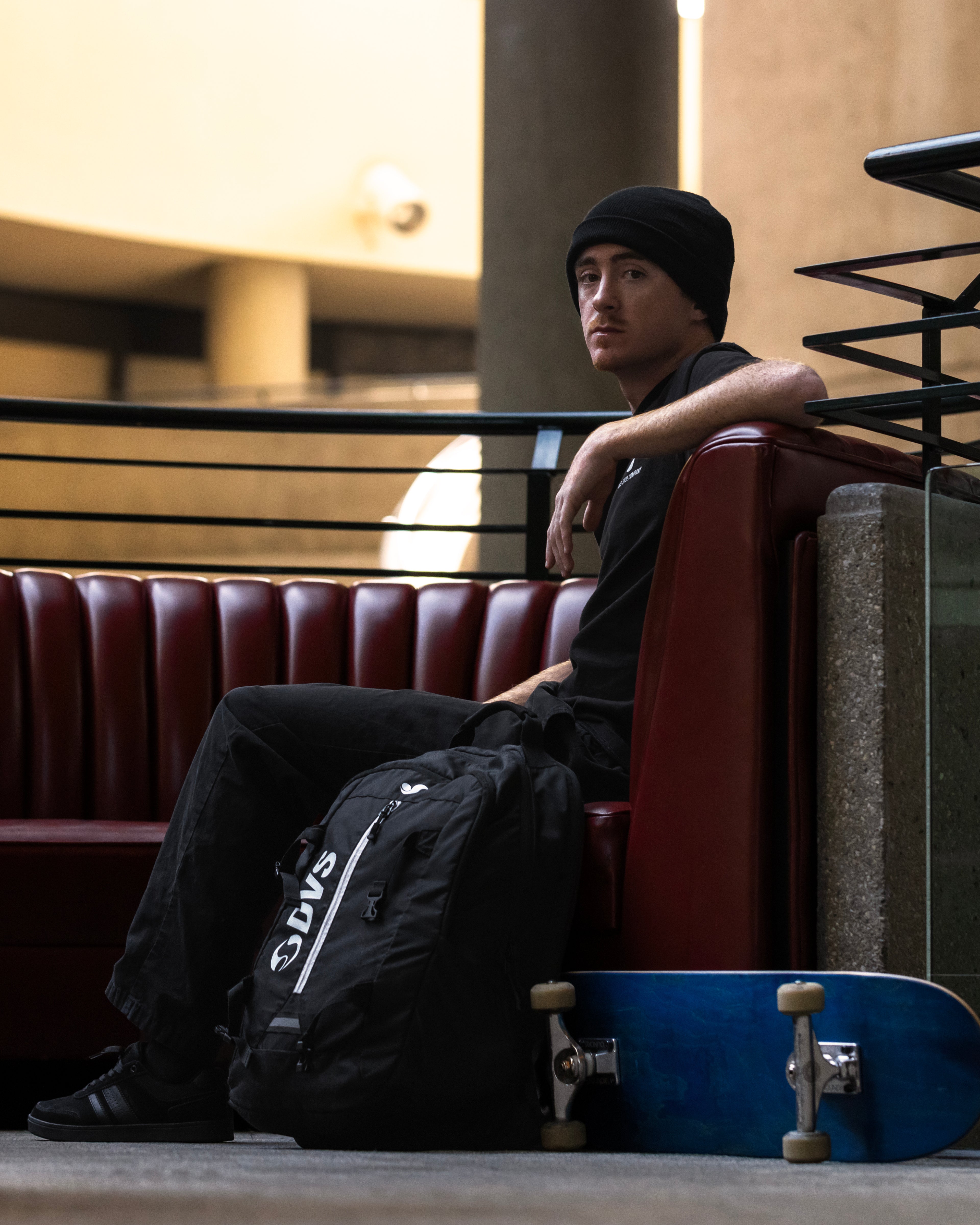 Person sitting on a red couch with a black backpack and blue skateboard in an indoor setting