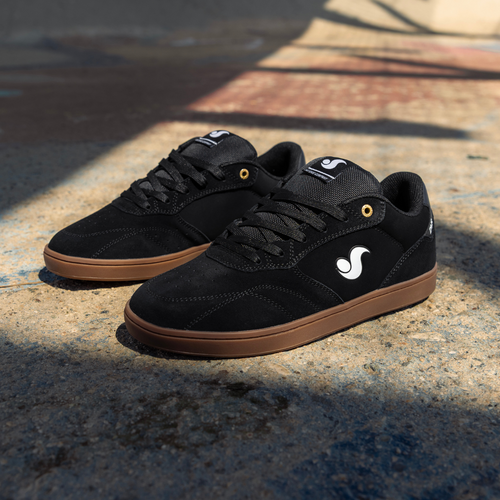 A pair of black skate shoes with brown gum soles and a white stylized wave logo on the side, displayed on a concrete surface in natural sunlight.